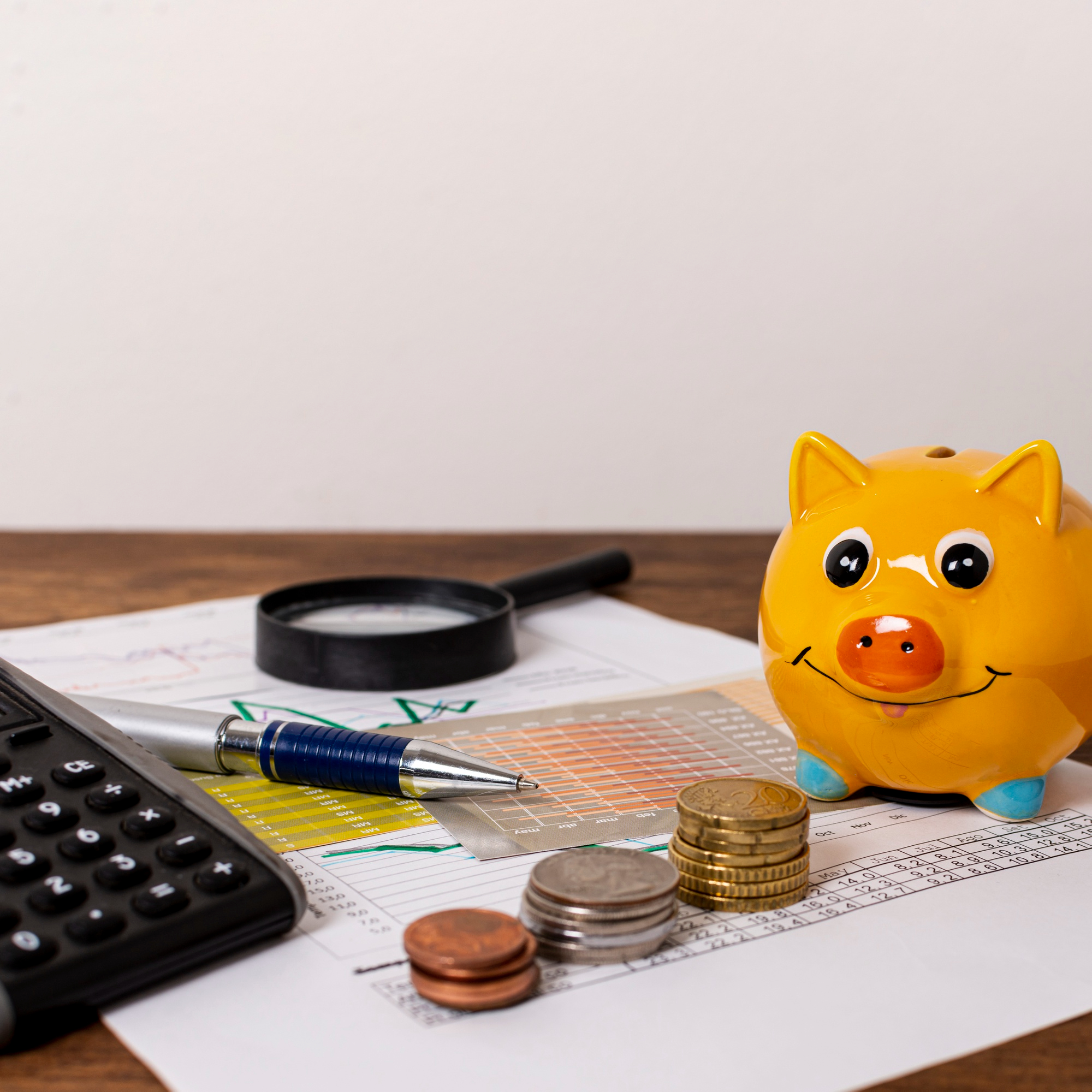 Estimation des aides et économies Tirelire jaune en forme de cochon posée sur des documents financiers, accompagnée de pièces de monnaie, d’un stylo et d’une loupe.