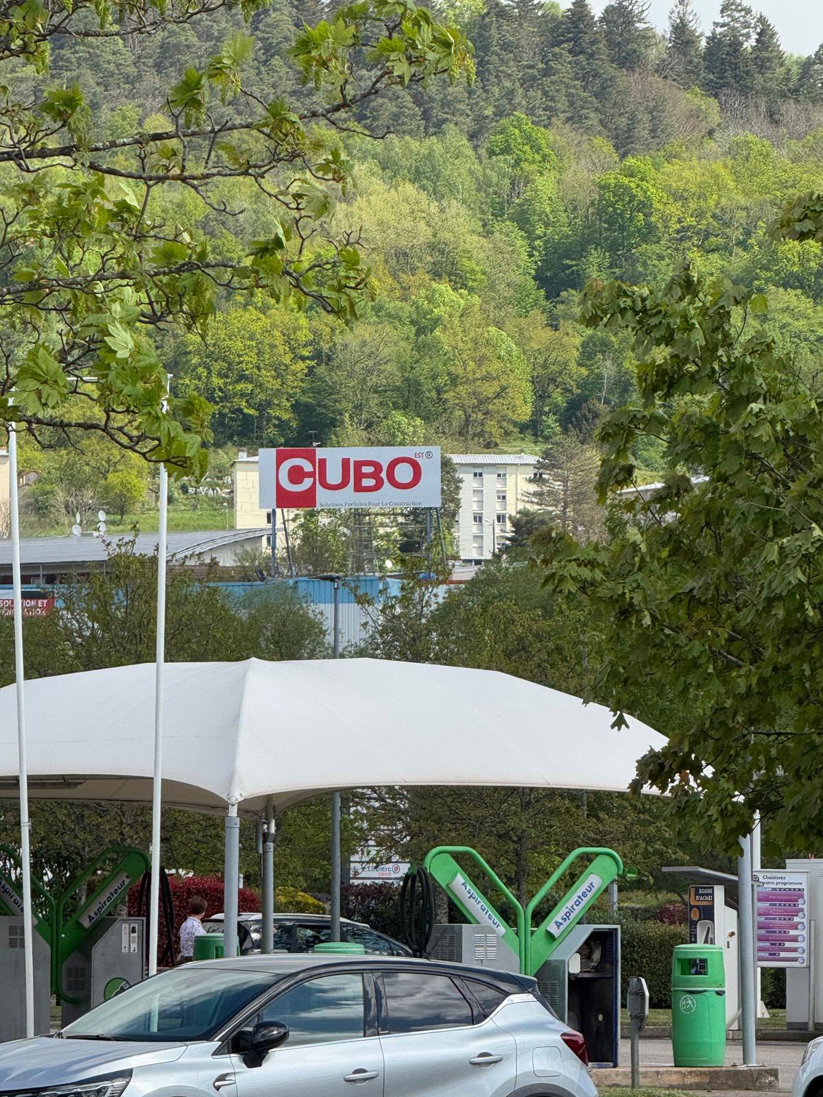 Siège de l’entreprise CUBO EST Vue du panneau d’entrée CUBO EST, visible depuis le parking, avec une station de lavage au premier plan et des arbres en arrière-plan.